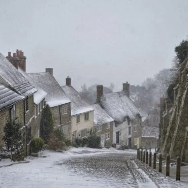 Snowy historic street: stone houses with snow-covered roofs on left, a ancient stone wall on right, light snowfall over cobbled road.