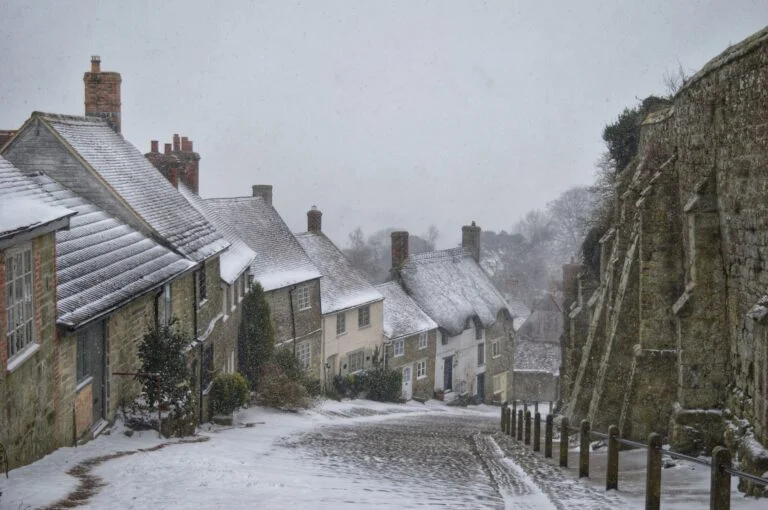Snowy historic street: stone houses with snow-covered roofs on left, a ancient stone wall on right, light snowfall over cobbled road.