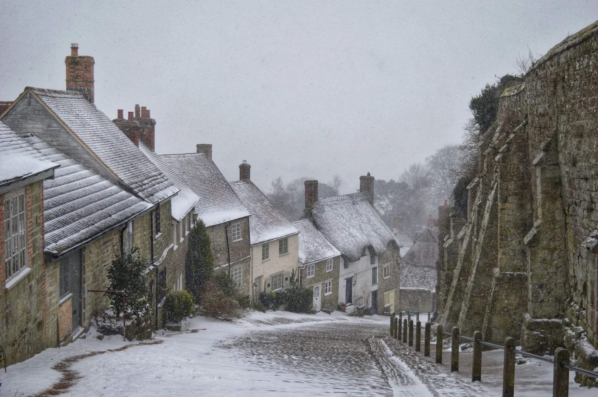 Snowy historic street: stone houses with snow-covered roofs on left, a ancient stone wall on right, light snowfall over cobbled road.