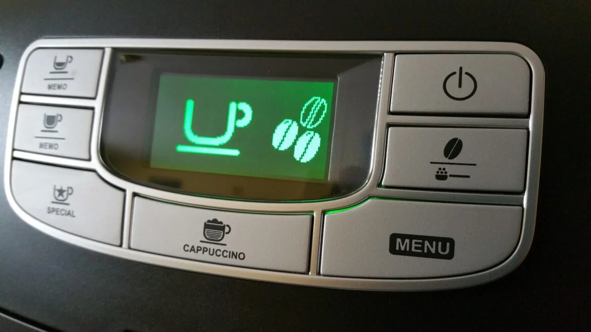 Close-up of a coffee machine control panel with a green LCD showing 'UP' and three coffee bean icons; surrounding buttons include Cappuccino, Menu, Power, and mug icons for beverages.