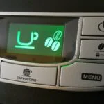 Close-up of a coffee machine control panel with a green LCD showing 'UP' and three coffee bean icons; surrounding buttons include Cappuccino, Menu, Power, and mug icons for beverages.