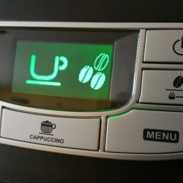 Close-up of a coffee machine control panel with a green LCD showing 'UP' and three coffee bean icons; surrounding buttons include Cappuccino, Menu, Power, and mug icons for beverages.