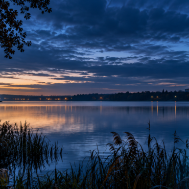 Evening lakeside scene with calm water, orange sunset on the horizon, dark blue clouds, and distant city lights reflecting on the lake.