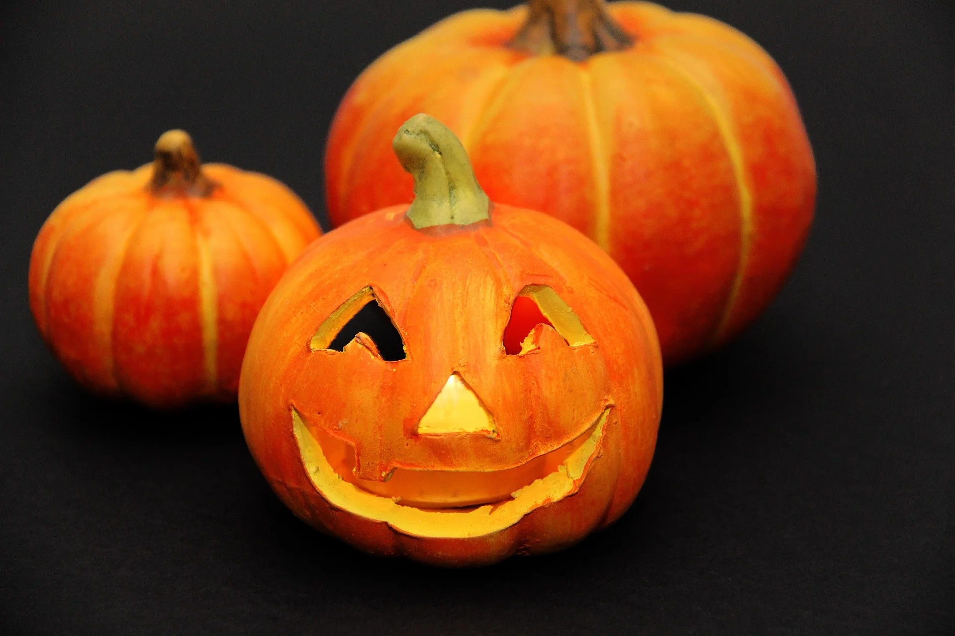 Three carved pumpkins sit on a dark surface, with the front pumpkin smiling and the others in the background.