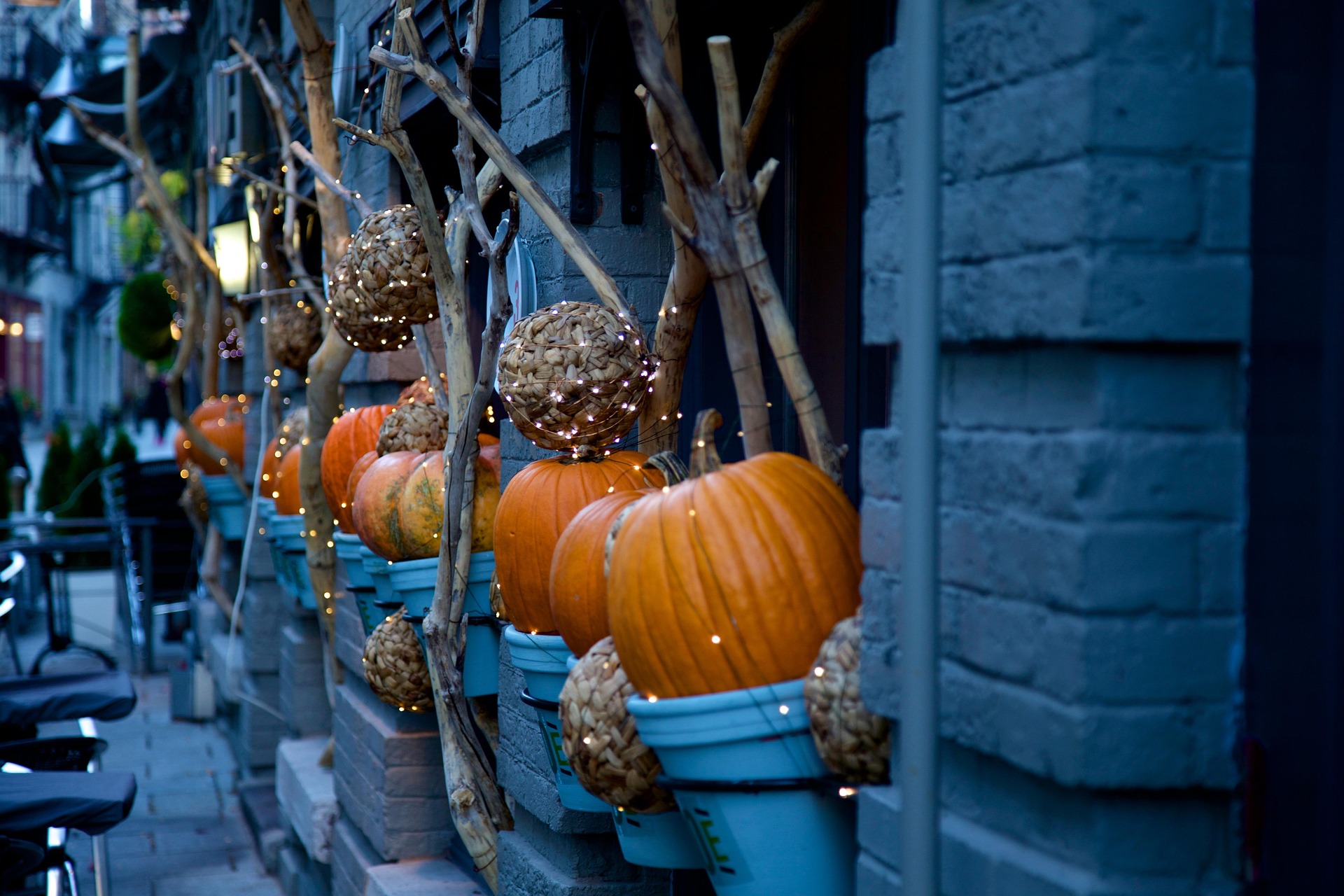 Row of pumpkins and woven wicker balls hung in blue planters along a brick storefront, lit by string lights.