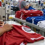 Worker sews a red sports jersey on a sewing machine in a garment factory, with folded shirts nearby on the table.