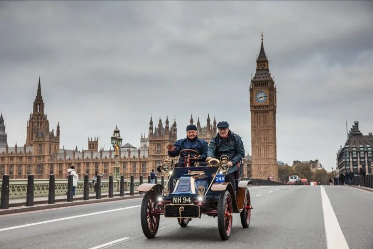 Two men in a vintage blue car drive across Westminster Bridge, with Big Ben and the Houses of Parliament in the background.