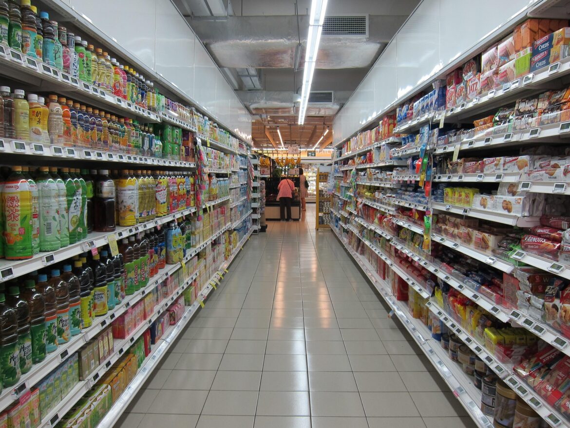 Aisle in a grocery store with tall shelves of beverages on both sides and two shoppers in the distance.