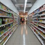 Aisle in a grocery store with tall shelves of beverages on both sides and two shoppers in the distance.