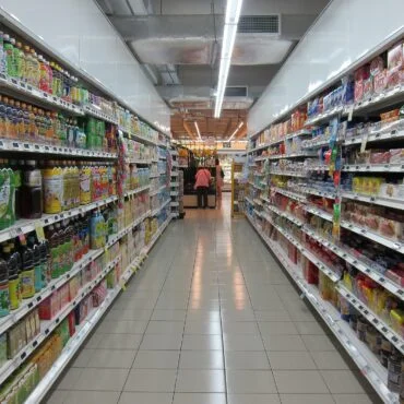 Aisle in a grocery store with tall shelves of beverages on both sides and two shoppers in the distance.