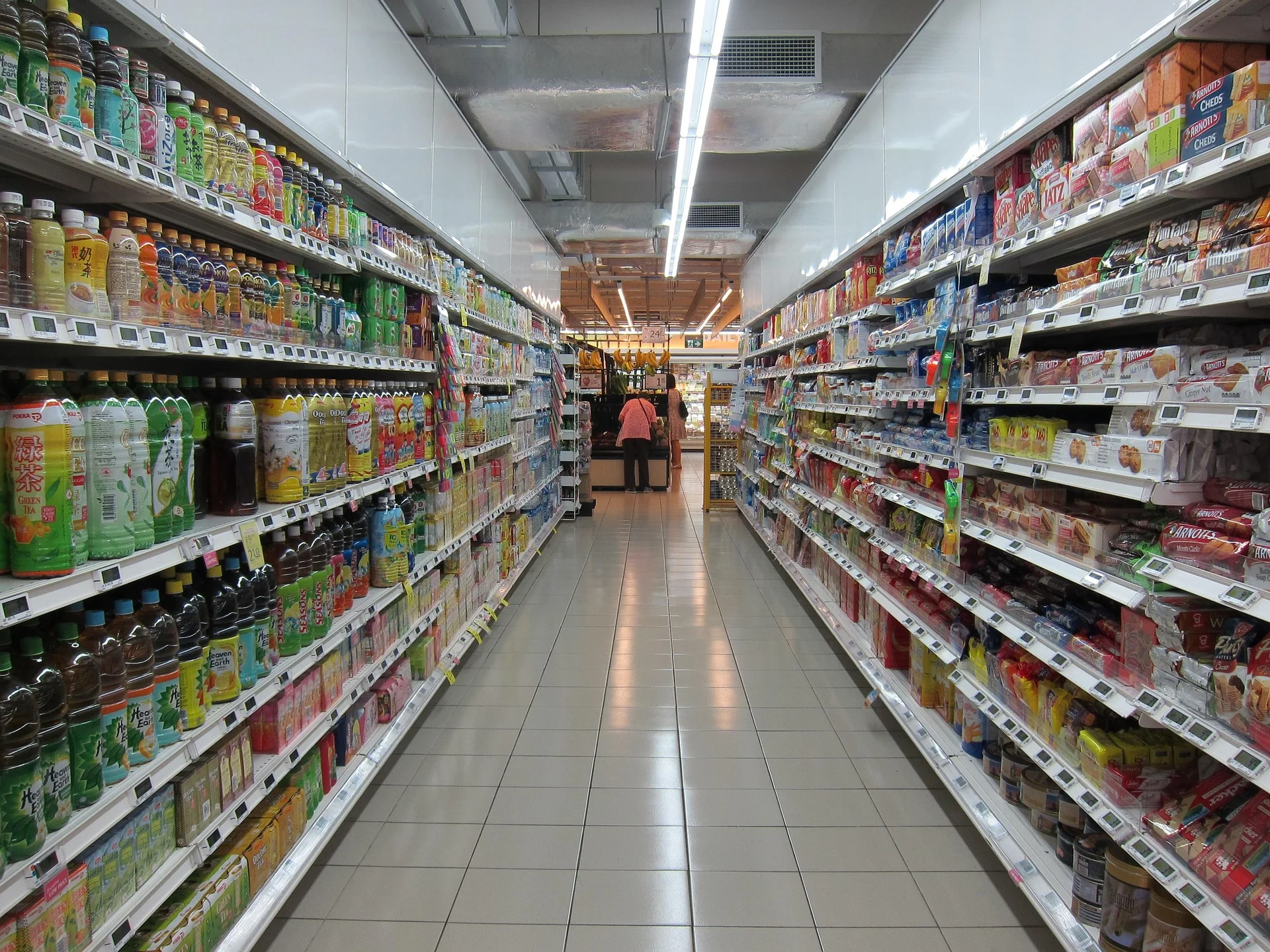 Aisle in a grocery store with tall shelves of beverages on both sides and two shoppers in the distance.