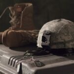 Camouflage military helmet with a front mount resting on a rugged storage case, with a brown combat boot and tangled cords nearby.