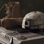 Camouflage military helmet with a front mount resting on a rugged storage case, with a brown combat boot and tangled cords nearby.