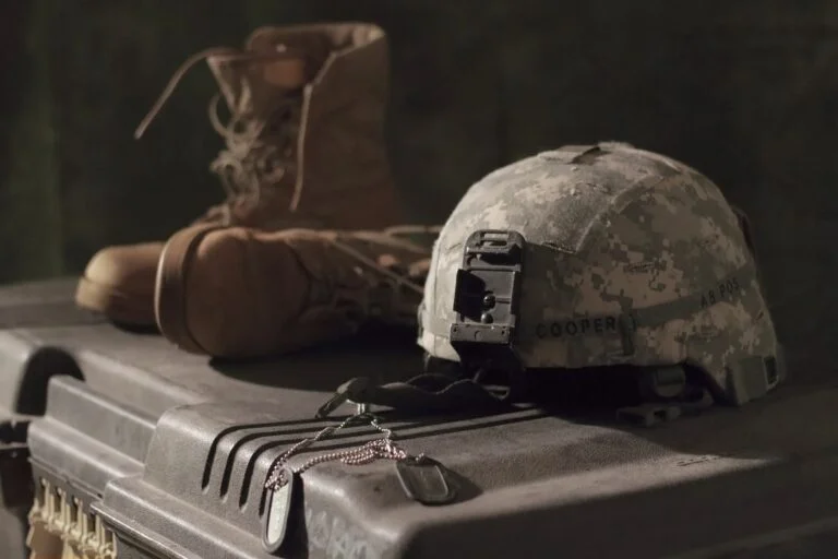 Camouflage military helmet with a front mount resting on a rugged storage case, with a brown combat boot and tangled cords nearby.