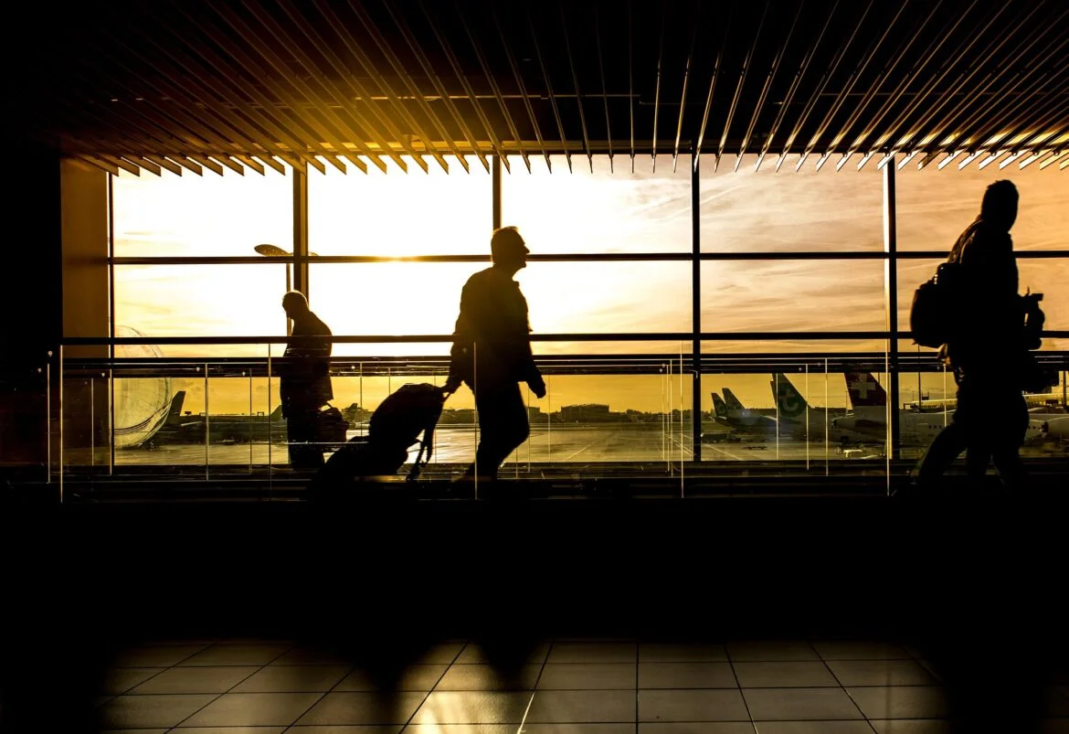Silhouetted travelers with luggage walk through an airport terminal at sunset, large windows and striped ceiling above.Outside, airplanes are visible on the tarmac.