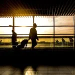 Silhouetted travelers with luggage walk through an airport terminal at sunset, large windows and striped ceiling above.Outside, airplanes are visible on the tarmac.
