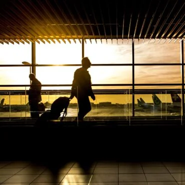 Silhouetted travelers with luggage walk through an airport terminal at sunset, large windows and striped ceiling above.Outside, airplanes are visible on the tarmac.