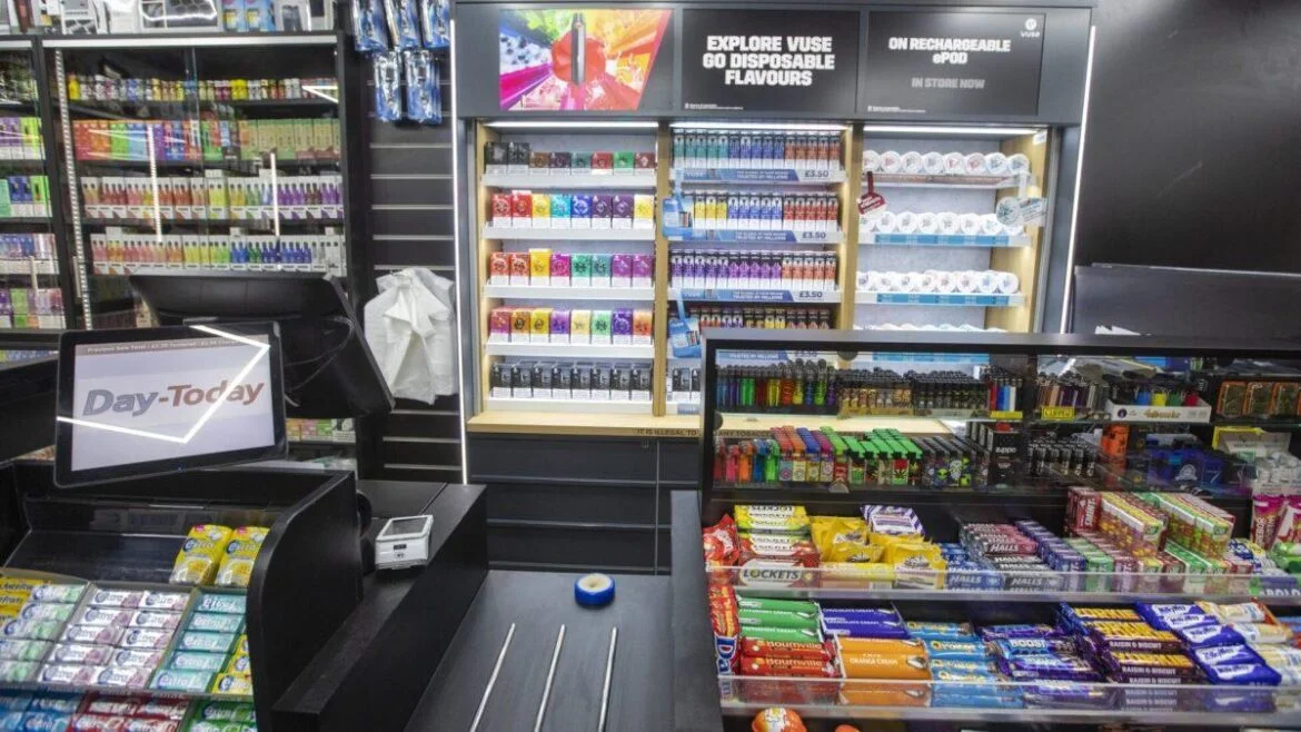 Checkout counter with a Day‑Today sign, surrounded by shelves of snacks, candy, and assorted tobacco/vape products in a store.