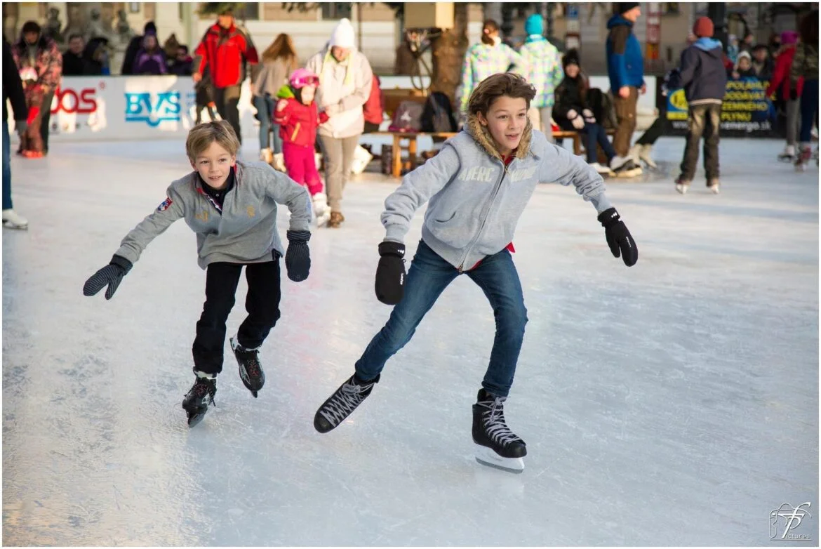 Two young boys skate on an outdoor ice rink, leaning forward as they glide, with spectators and skaters in the background.