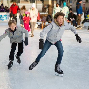 Two young boys skate on an outdoor ice rink, leaning forward as they glide, with spectators and skaters in the background.