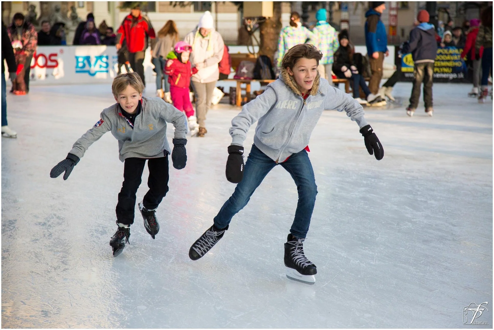 Two young boys skate on an outdoor ice rink, leaning forward as they glide, with spectators and skaters in the background.