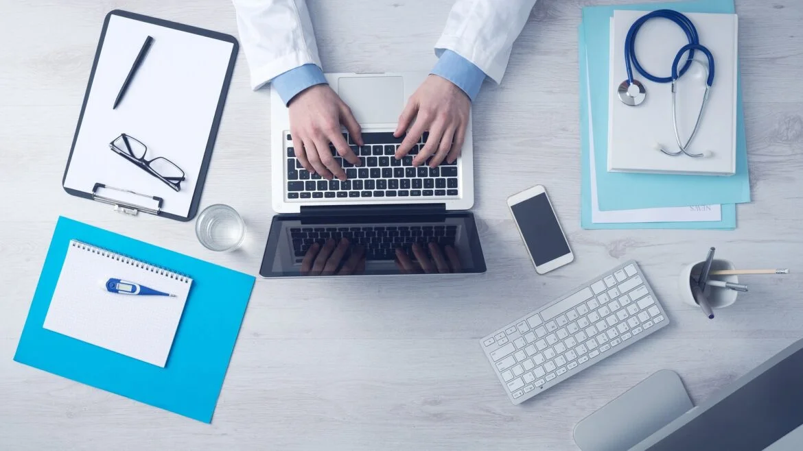 Top-down view of someone in a white coat typing on a laptop at a clinic desk, with a stethoscope and documents nearby.