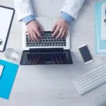 Top-down view of someone in a white coat typing on a laptop at a clinic desk, with a stethoscope and documents nearby.