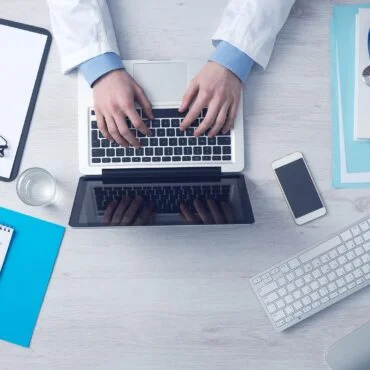 Top-down view of someone in a white coat typing on a laptop at a clinic desk, with a stethoscope and documents nearby.