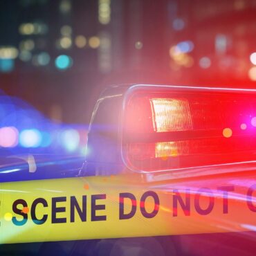 Police car with red and blue lights behind yellow 'CRIME SCENE DO NOT CROSS' tape at night city scene.