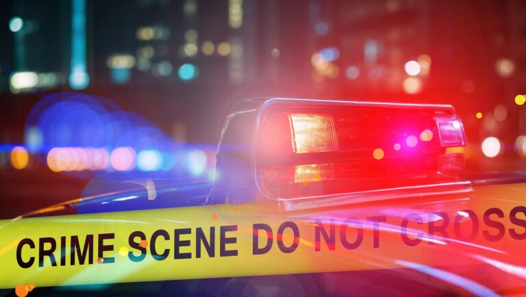 Police car with red and blue lights behind yellow 'CRIME SCENE DO NOT CROSS' tape at night city scene.