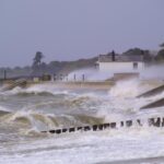 Waves crash over a seawall at a harbor, with a small white building in the distance and wind-swept trees beyond.
