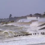 Rough coastal scene with large waves crashing over a seawall toward a small white house in the distance.