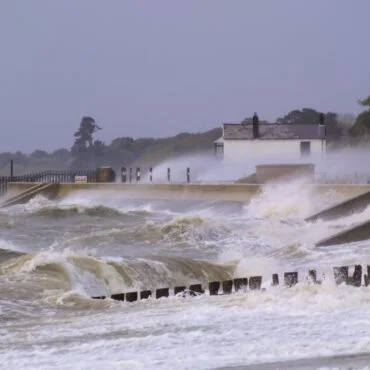 Rough coastal scene with large waves crashing over a seawall toward a small white house in the distance.