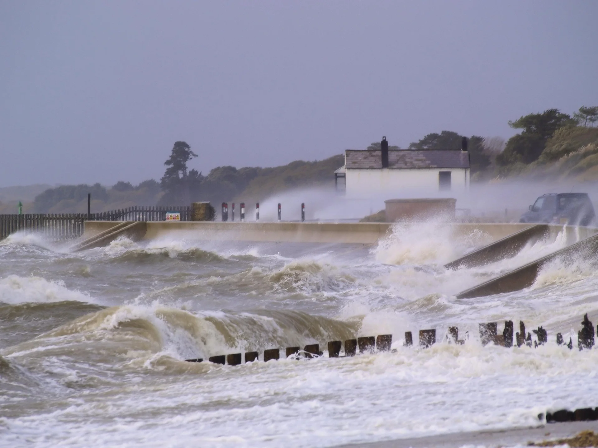 Rough coastal scene with large waves crashing over a seawall toward a small white house in the distance.