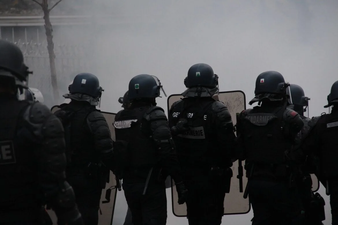 Line of riot police in black armor with helmets and shields, facing forward in a smoky scene.