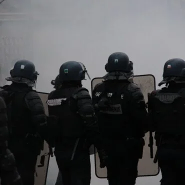 Line of riot police in black armor with helmets and shields, facing forward in a smoky scene.