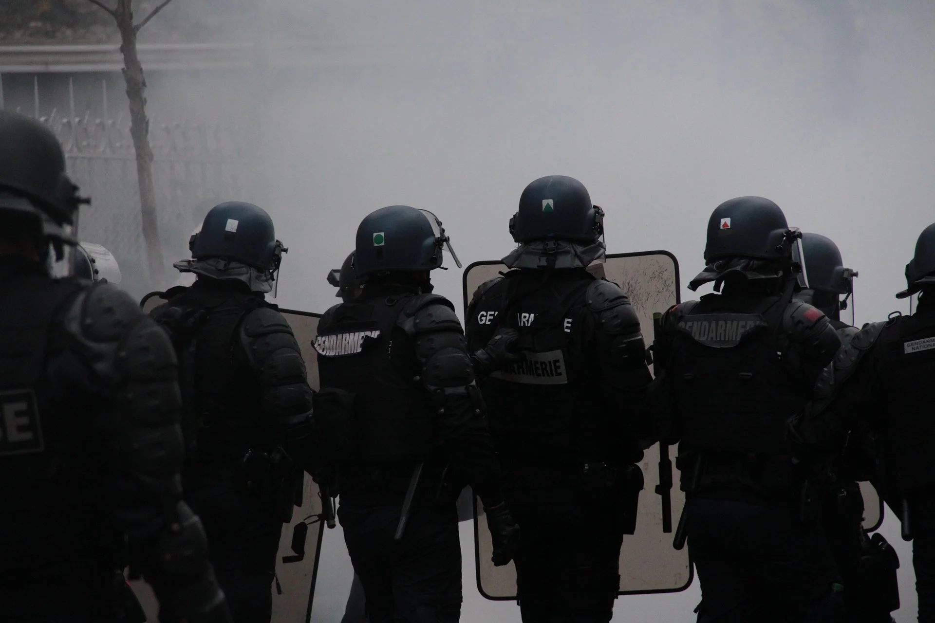 Line of riot police in black armor with helmets and shields, facing forward in a smoky scene.