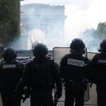 Riot police in full armor with shields standing on a smoky Paris street; Arc de Triomphe visible in the background.