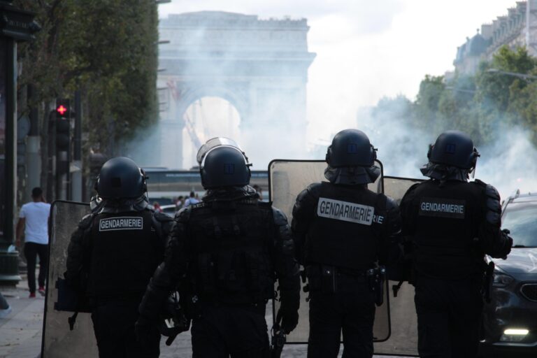 Riot police in full armor with shields standing on a smoky Paris street; Arc de Triomphe visible in the background.
