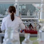 Back view of a scientist in a white lab coat working at a bench filled with glass bottles and lab equipment in a laboratory stage setting.