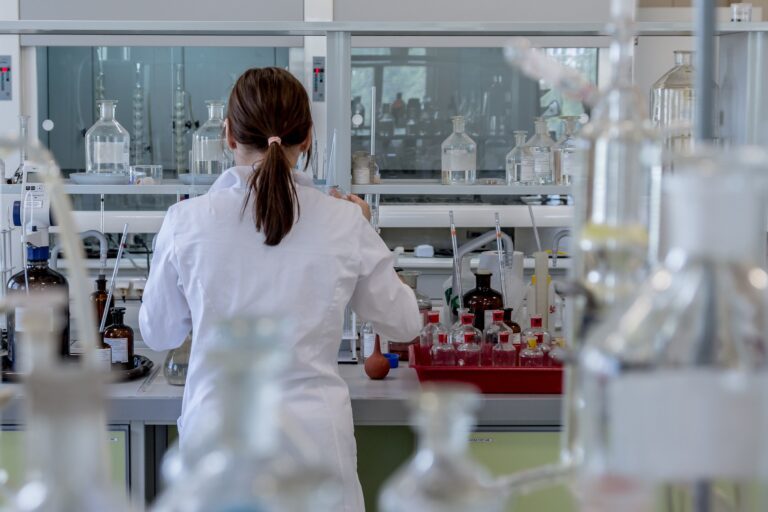 Back view of a scientist in a white lab coat working at a bench filled with glass bottles and lab equipment in a laboratory stage setting.