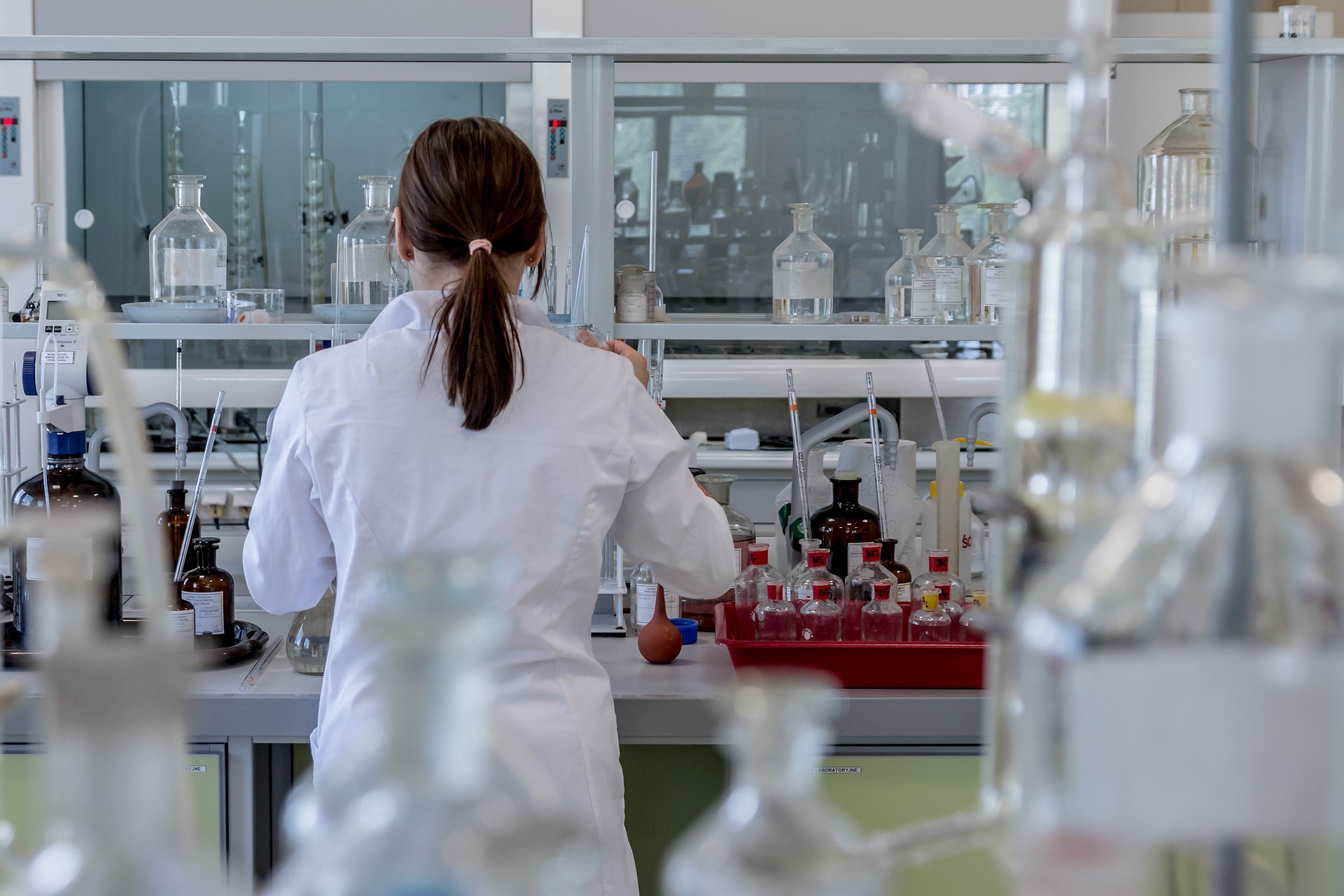 Back view of a scientist in a white lab coat working at a bench filled with glass bottles and lab equipment in a laboratory stage setting.