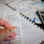Person writes with an orange pencil over a receipt and a spiral-bound notebook with grid paper, calculator nearby, suggesting budgeting or expenses tracking.