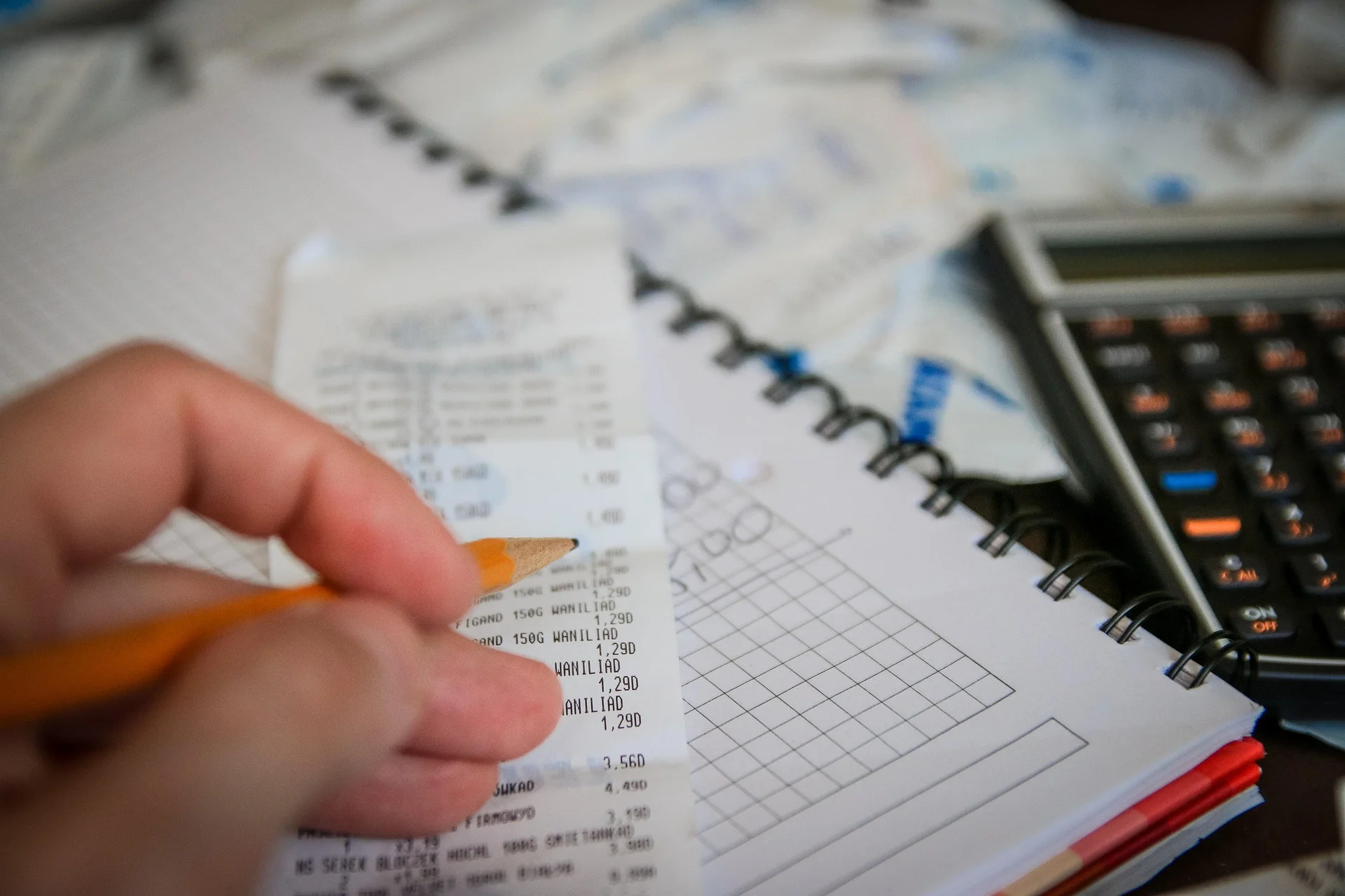 Person writes with an orange pencil over a receipt and a spiral-bound notebook with grid paper, calculator nearby, suggesting budgeting or expenses tracking.
