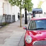 Red compact car parked along a city sidewalk, featuring a Union Jack-pattern side mirror and reflections on the windshield in a sunny urban street setting