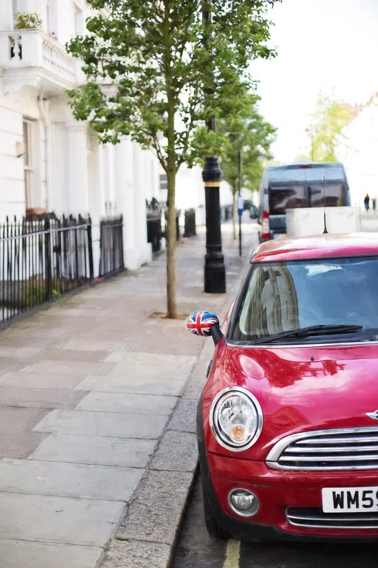 Red compact car parked along a city sidewalk, featuring a Union Jack-pattern side mirror and reflections on the windshield in a sunny urban street setting