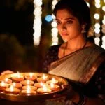 Indian woman in a saree holding a brass plate with lit oil lamps during a Diwali celebration at night, with warm bokeh lights in the background.