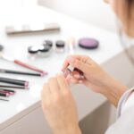 Close-up of hands applying pink nail polish at a white makeup table with brushes and cosmetics nearby on a bright vanity surface