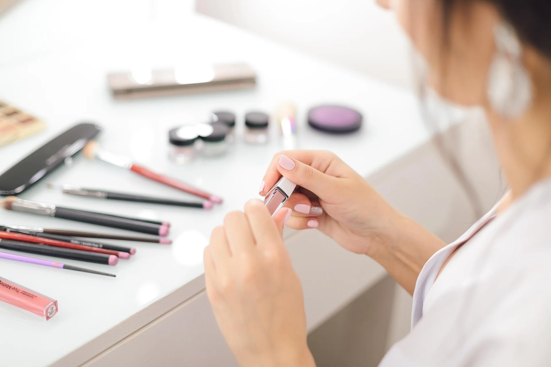 Close-up of hands applying pink nail polish at a white makeup table with brushes and cosmetics nearby on a bright vanity surface