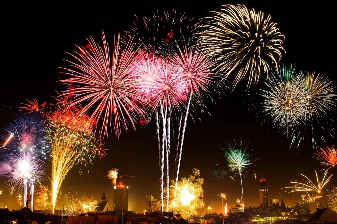 Colorful fireworks bursting over a city skyline at night, with red, pink, and gold trails lighting the sky.